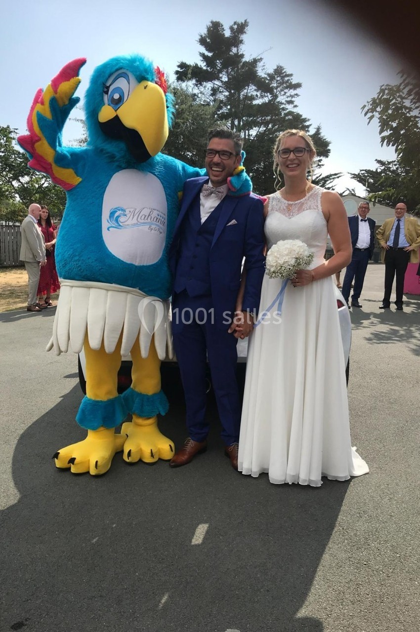 Un couple en tenue de mariage pose avec une mascotte colorée devant une voiture, en extérieur.