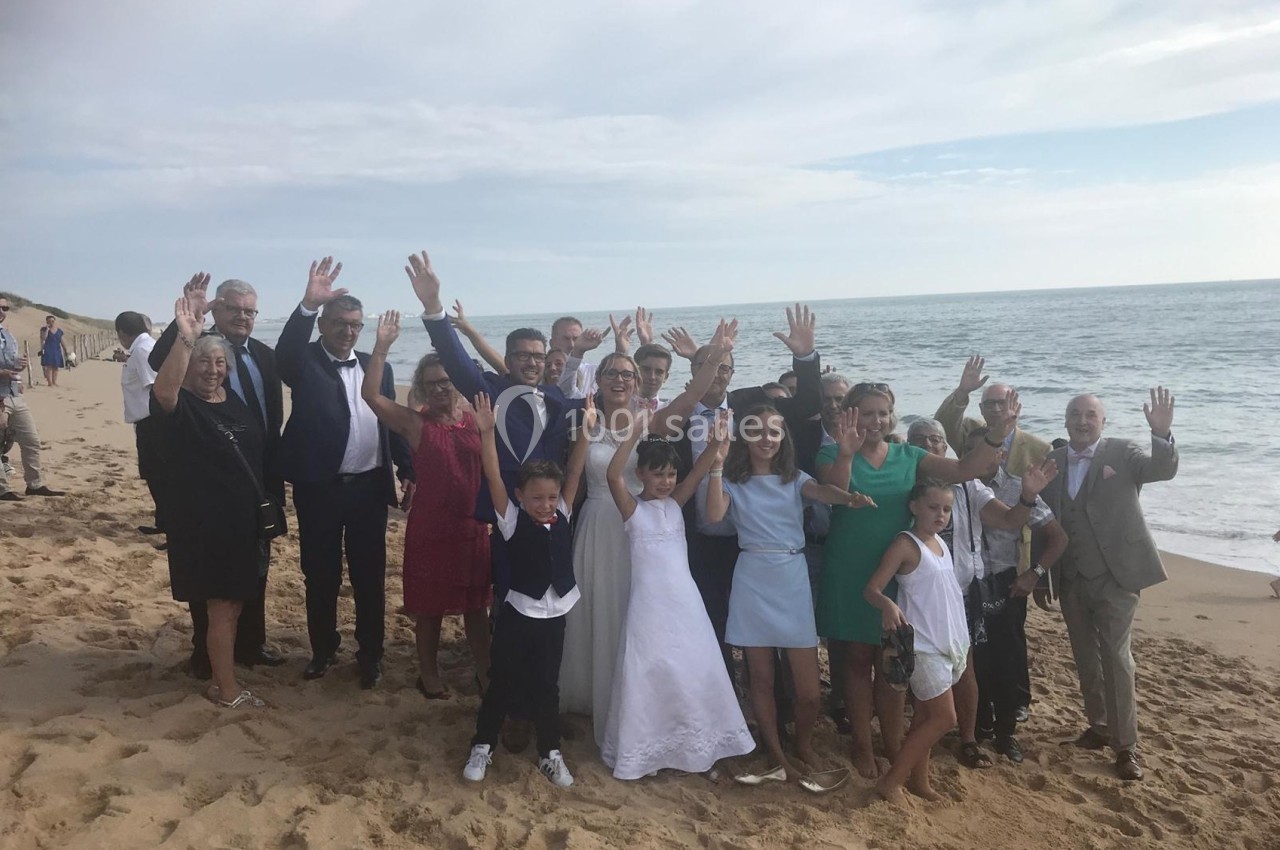 Un groupe de personnes souriantes pose sur une plage, levant les bras devant la mer sous un ciel partiellement nuageux.