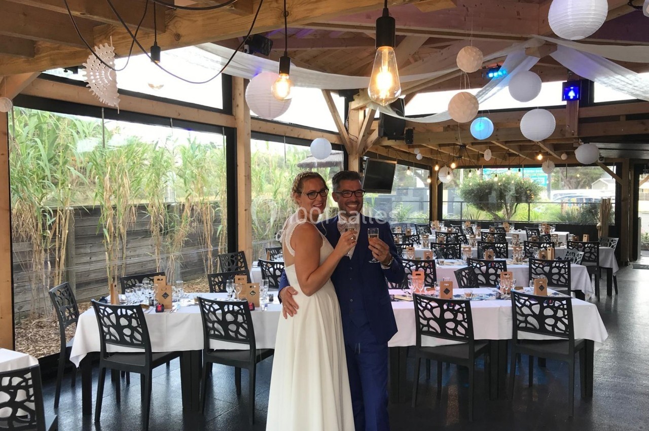 Un couple en tenue de mariage trinque dans une salle décorée avec des tables dressées et des lanternes suspendues.