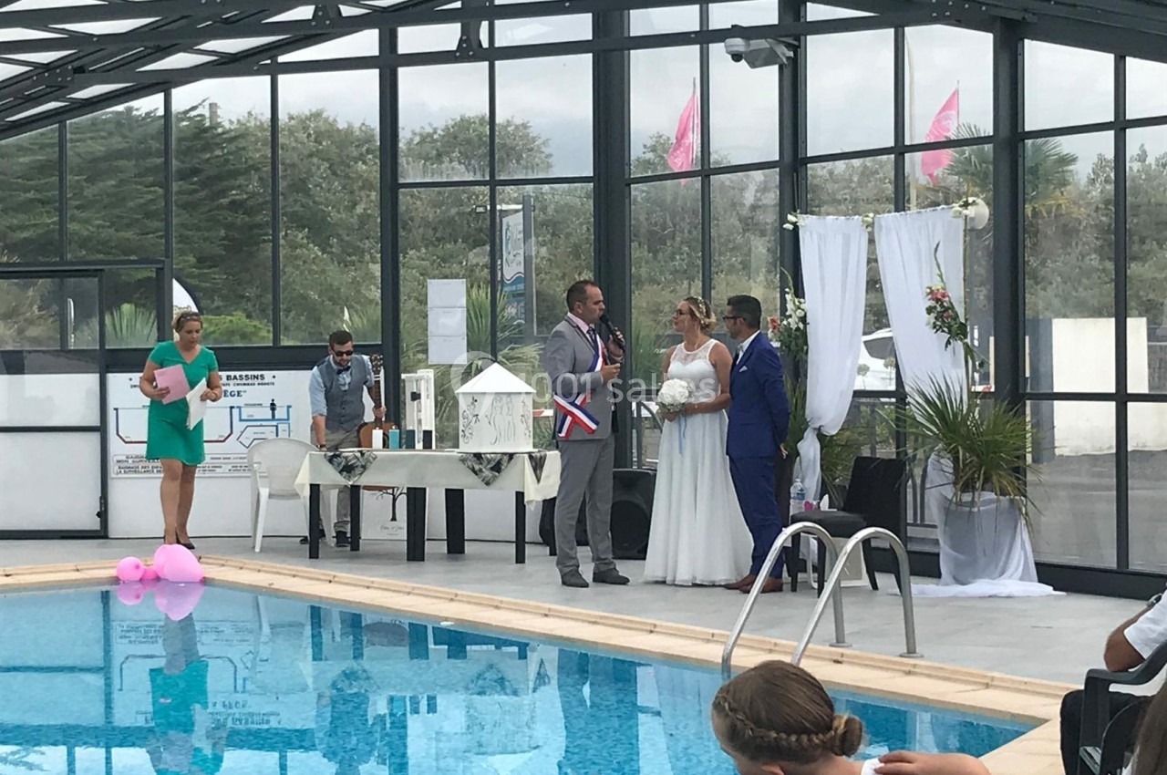 Un mariage célébré au bord d'une piscine couverte, avec un couple et un officiant devant un décor floral.