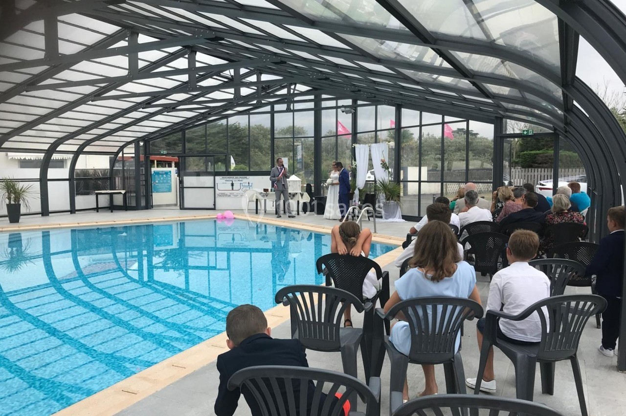Cérémonie près d'une piscine couverte, avec des invités assis et un couple debout devant une table décorée.