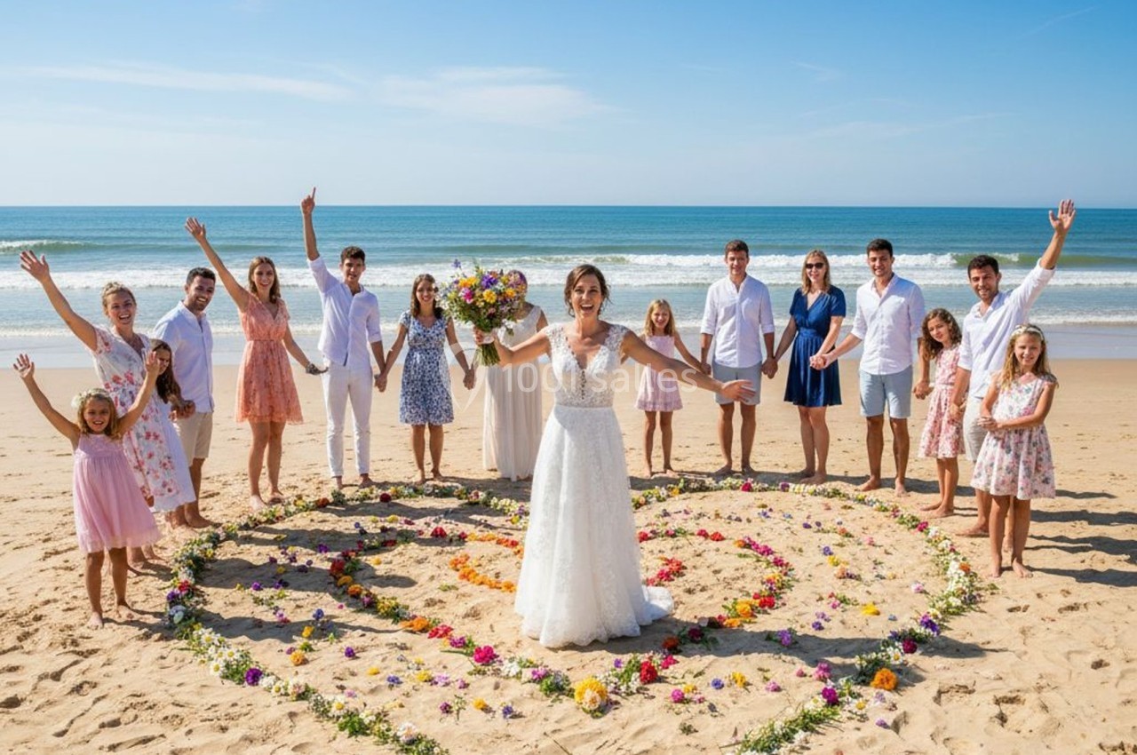 Un groupe de personnes souriantes entourant une mariée, debout sur une plage, dans un cœur formé de fleurs colorées.