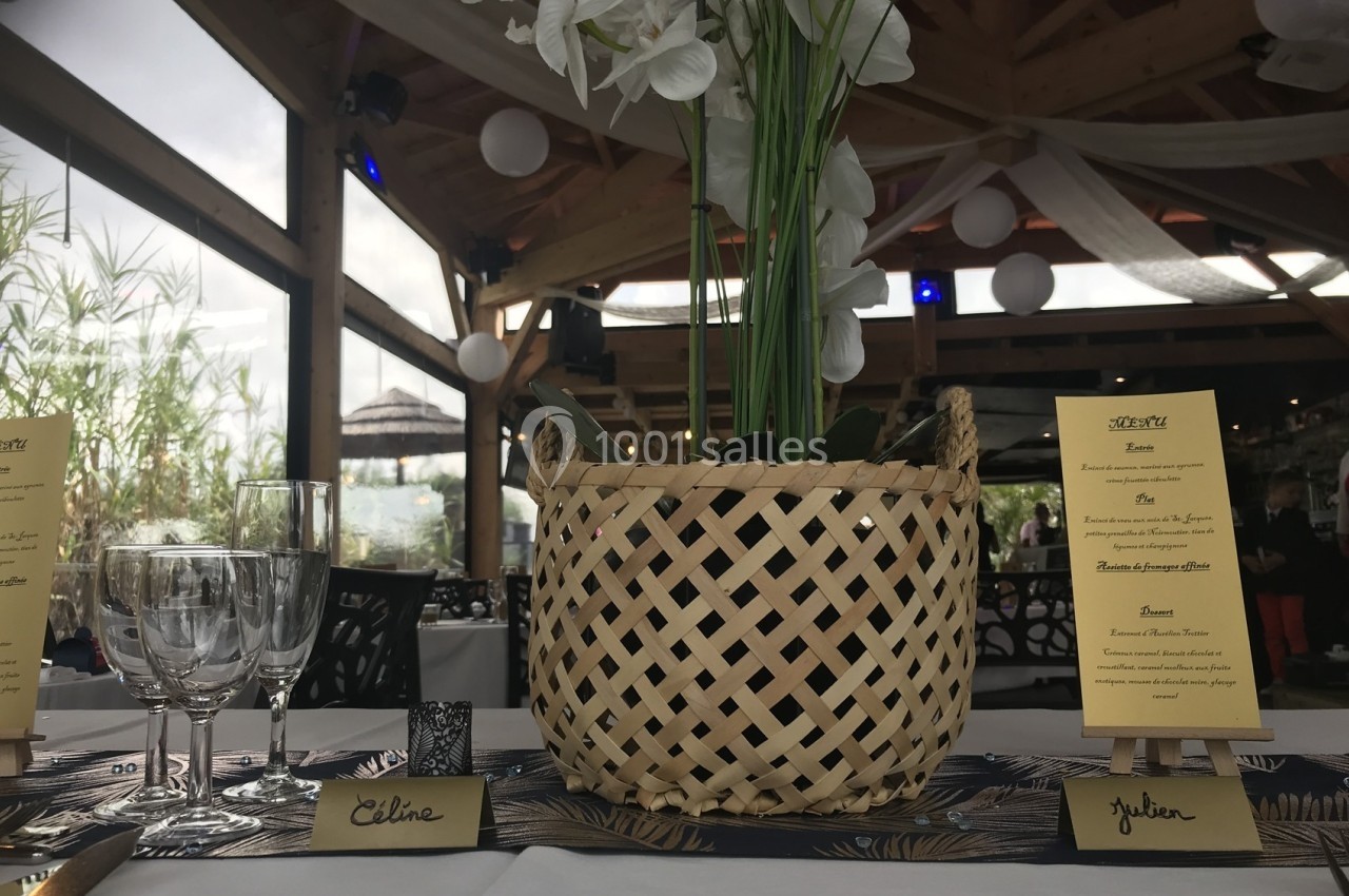 Centre de table avec une plante dans un panier en osier, entouré de menus et de verres sur une table dressée.