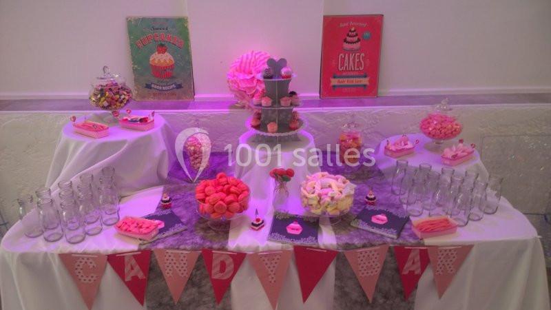 Table décorée pour une fête avec bonbons, gâteaux, bouteilles en verre et guirlande rose en avant-plan.