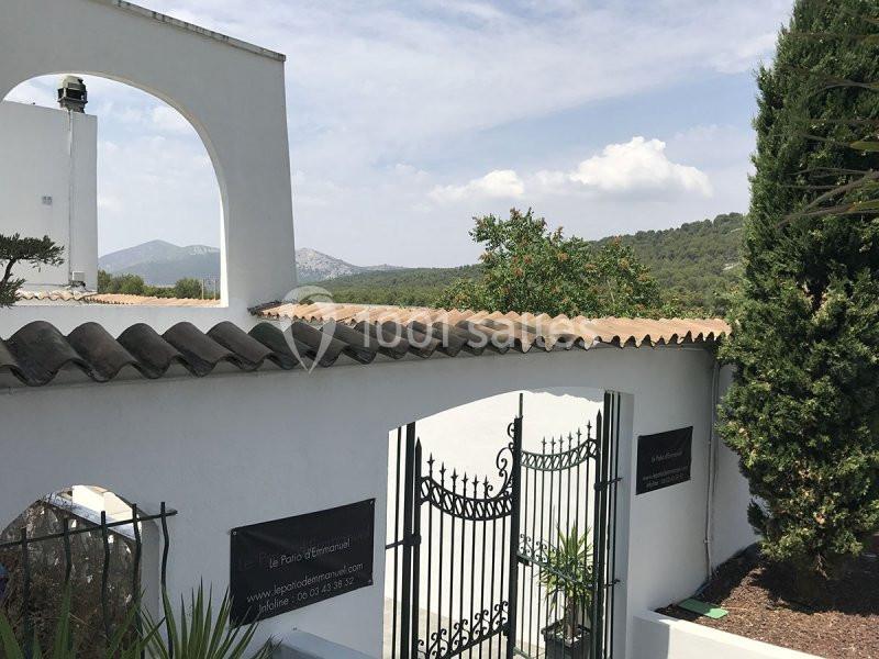Portail en fer forgé noir entouré de murs blancs, avec vue sur des collines boisées sous un ciel partiellement nuageux.