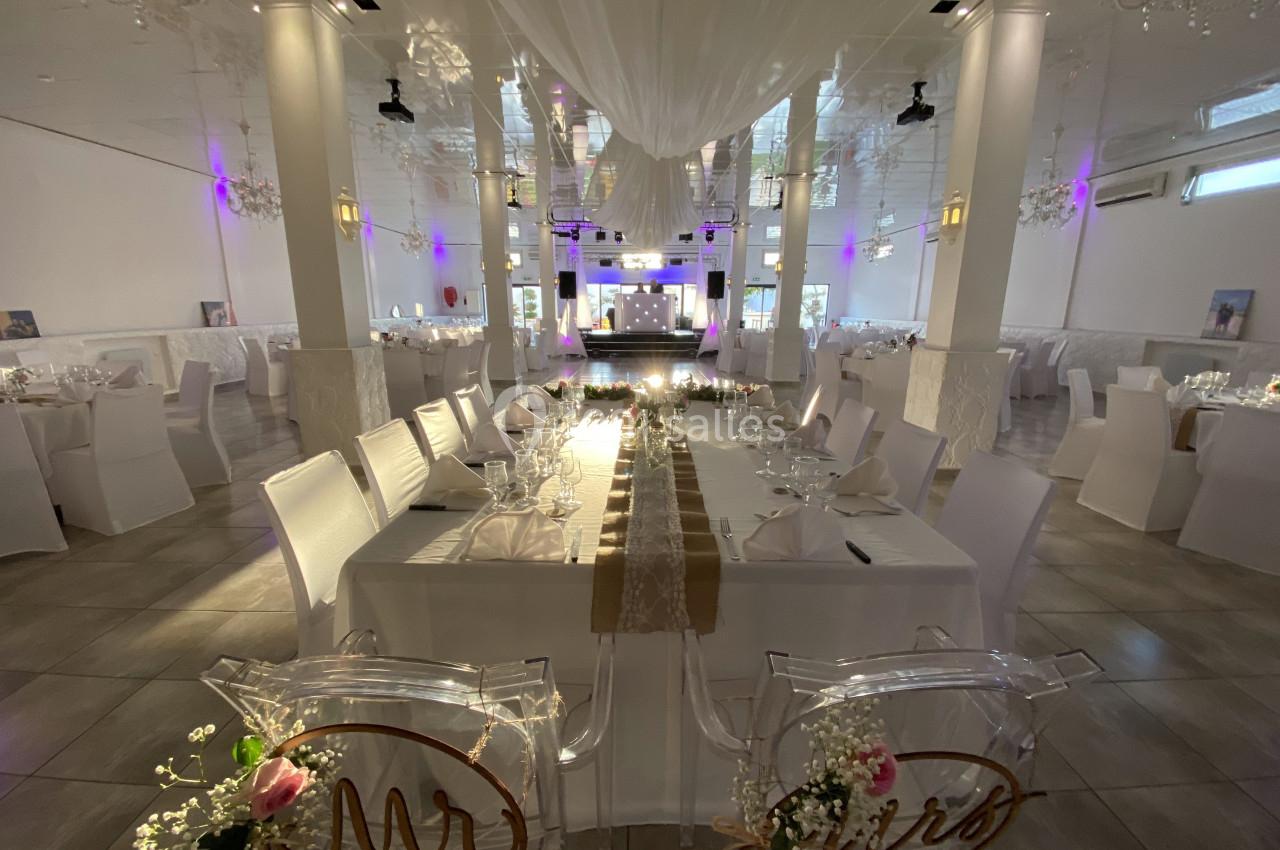 Salle de réception élégante avec tables décorées, chaises blanches et éclairage tamisé dans une ambiance festive.
