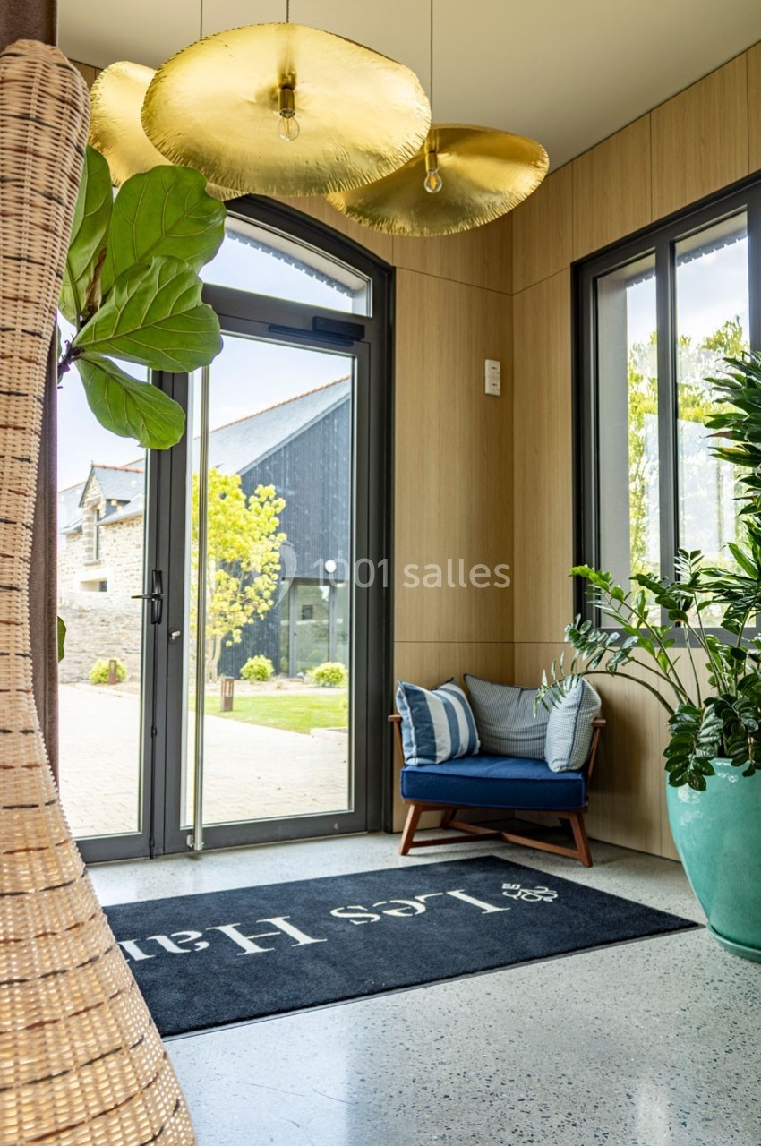 Entrée lumineuse avec porte vitrée, tapis noir, fauteuil bleu, plantes vertes et suspensions dorées.