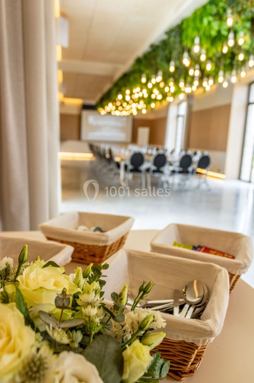 Salle de réunion lumineuse avec des tables disposées en U, décorée de plantes suspendues et d'un bouquet au premier plan.