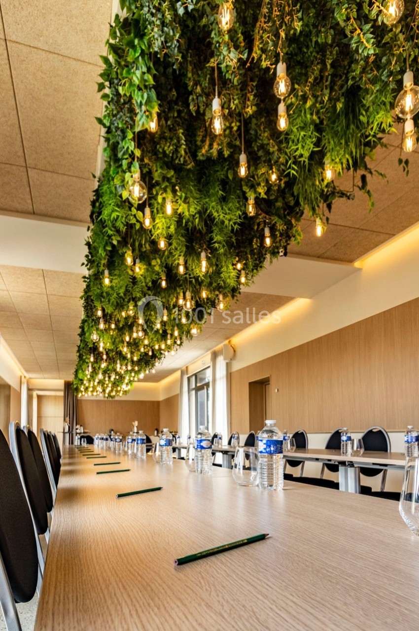 Salle de réunion avec une longue table en bois, des chaises noires et un plafond décoré de plantes et de lumières suspendues.