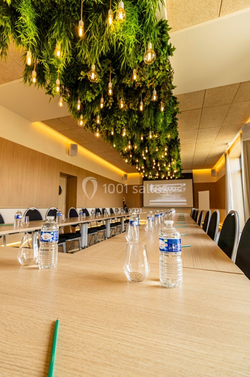 Salle de réunion moderne avec plafond végétalisé, éclairage suspendu et tables équipées de bouteilles d'eau et stylos.