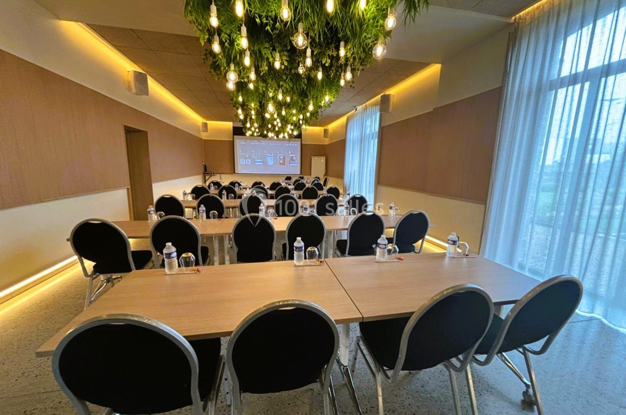 Salle de réunion lumineuse avec tables en bois, chaises noires, écran de présentation et suspension végétale.