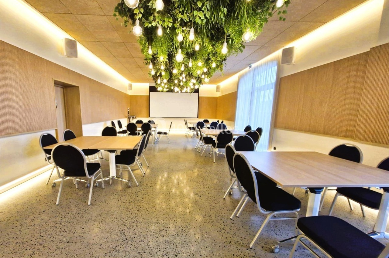Salle de réunion lumineuse avec tables rectangulaires, chaises noires et plafond décoré de plantes et ampoules suspendues.