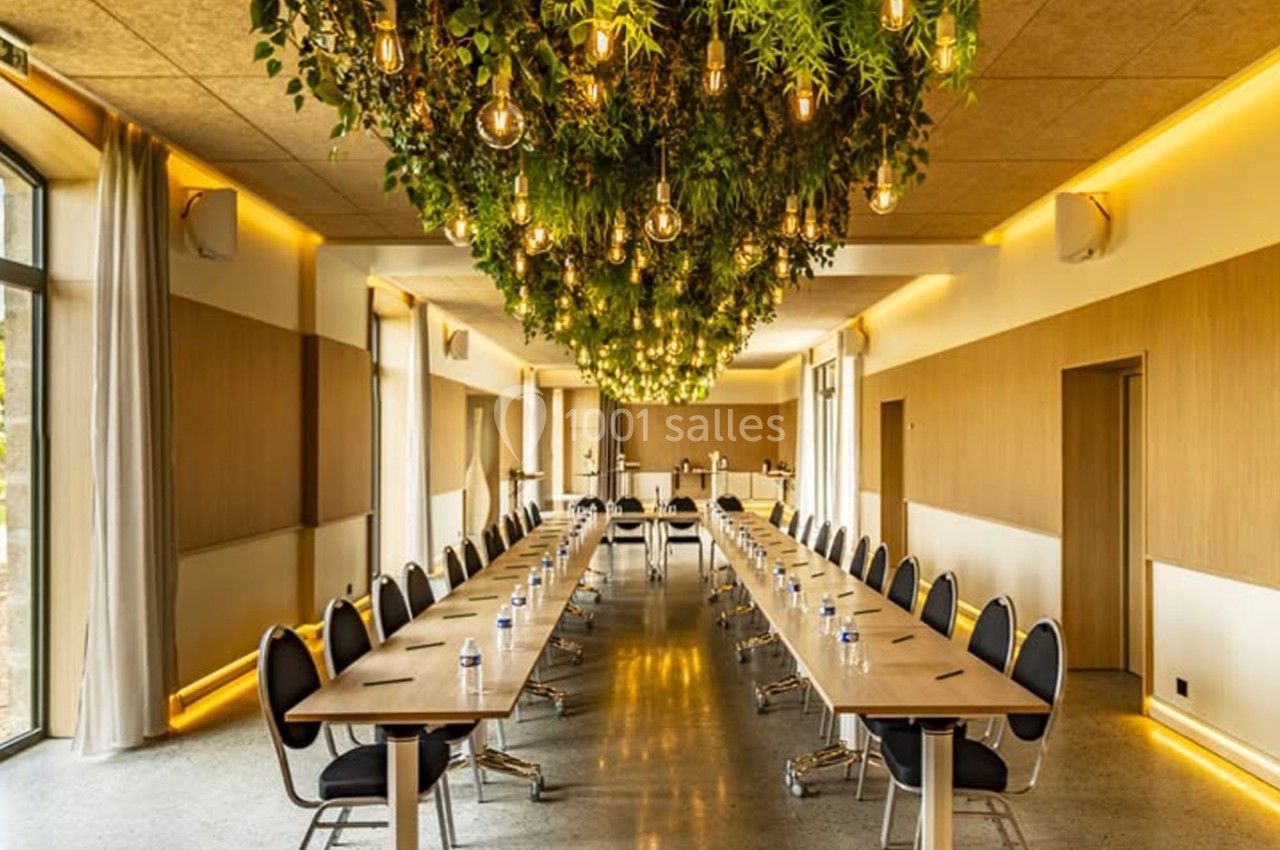 Salle de réunion lumineuse avec une longue table en U, des chaises noires et un plafond décoré de plantes suspendues.