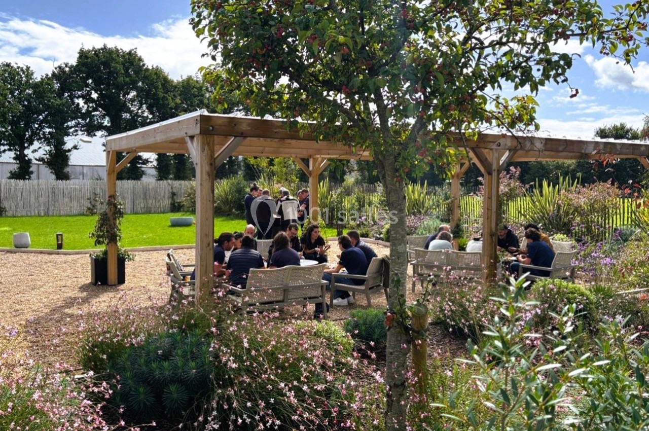 Un groupe de personnes assises sous une pergola en bois dans un jardin fleuri par une journée ensoleillée.