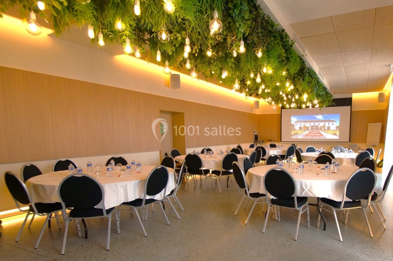 Salle de réunion lumineuse avec tables rondes dressées, plafond végétalisé et écran de projection en arrière-plan.
