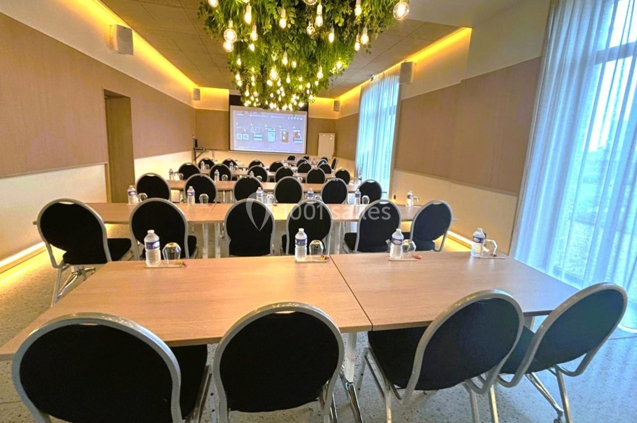 Salle de réunion lumineuse avec tables alignées, chaises noires, écran de projection et décoration végétale suspendue.