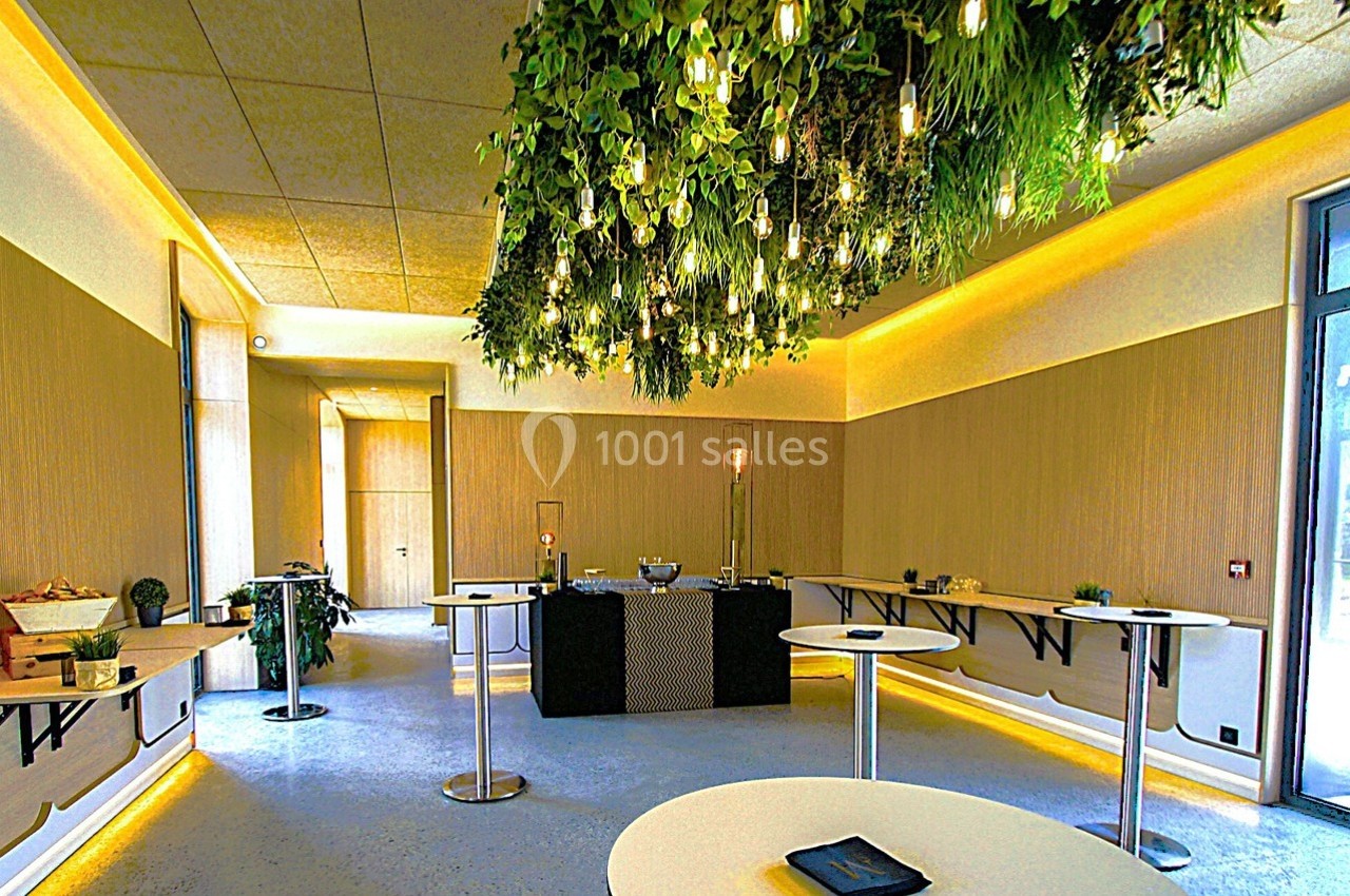 Salle lumineuse avec tables hautes, décor végétal suspendu et éclairage chaleureux.