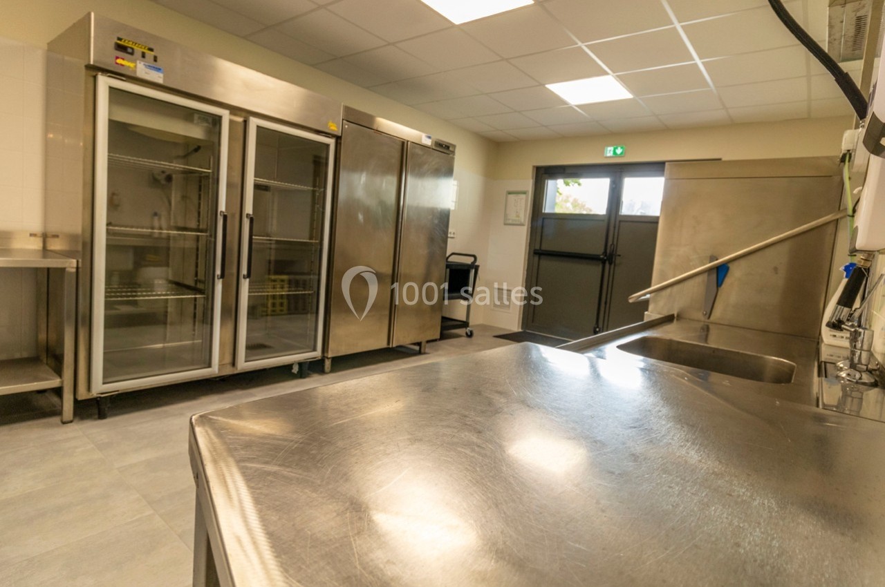 Cuisine professionnelle avec plans de travail en inox, réfrigérateurs industriels et évier sous un éclairage fluorescent.