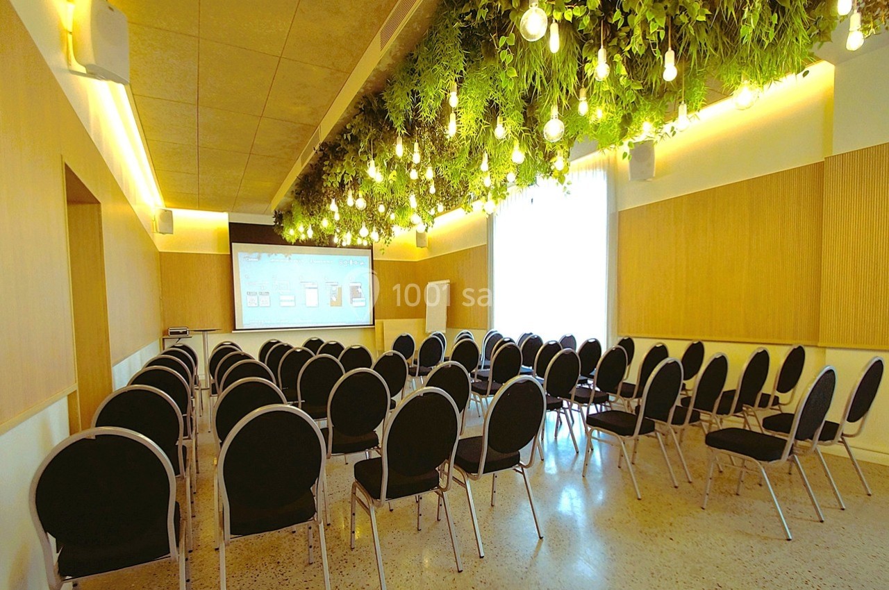 Salle de conférence moderne avec des chaises noires alignées, un écran de projection et un plafond végétalisé éclairé.