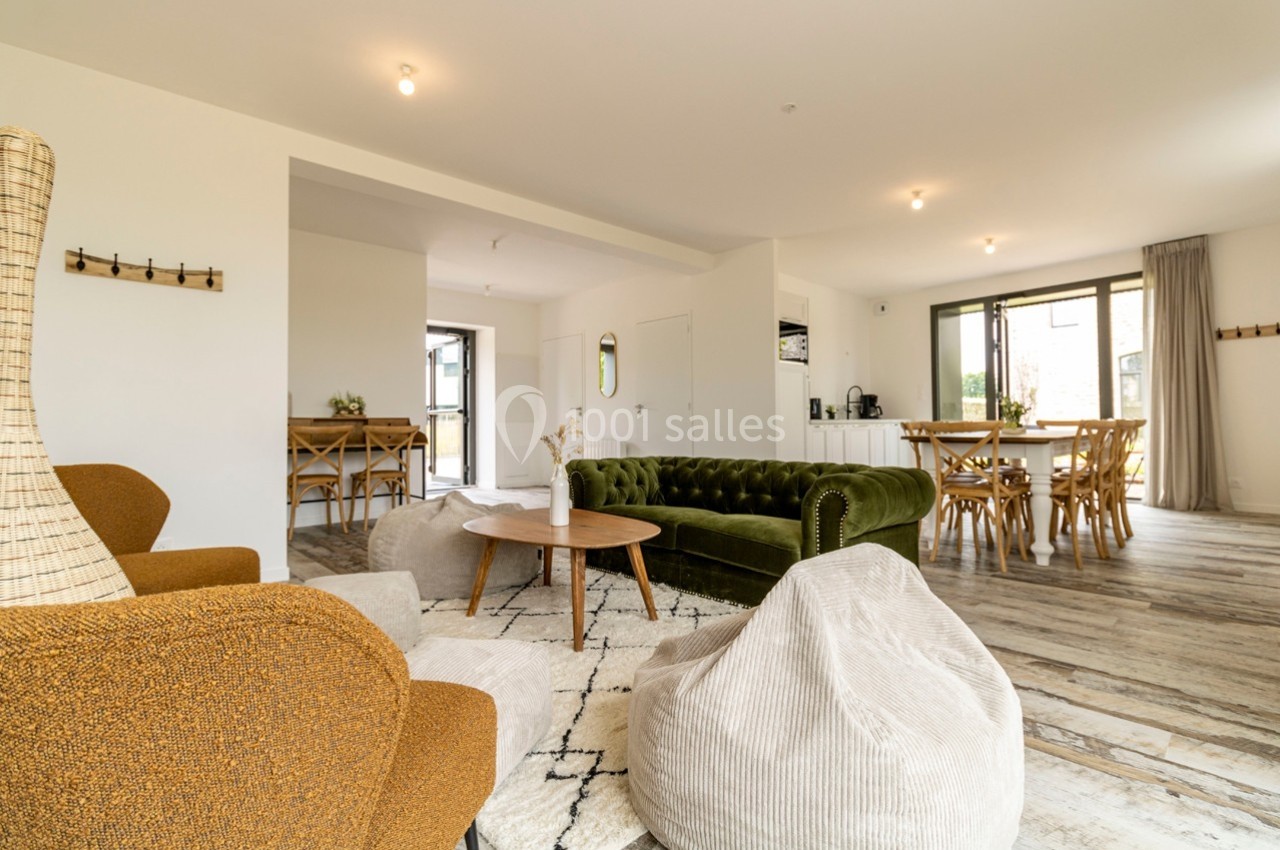 Salon spacieux avec canapé vert, fauteuils, poufs, table basse et coin repas, éclairé par de grandes fenêtres.