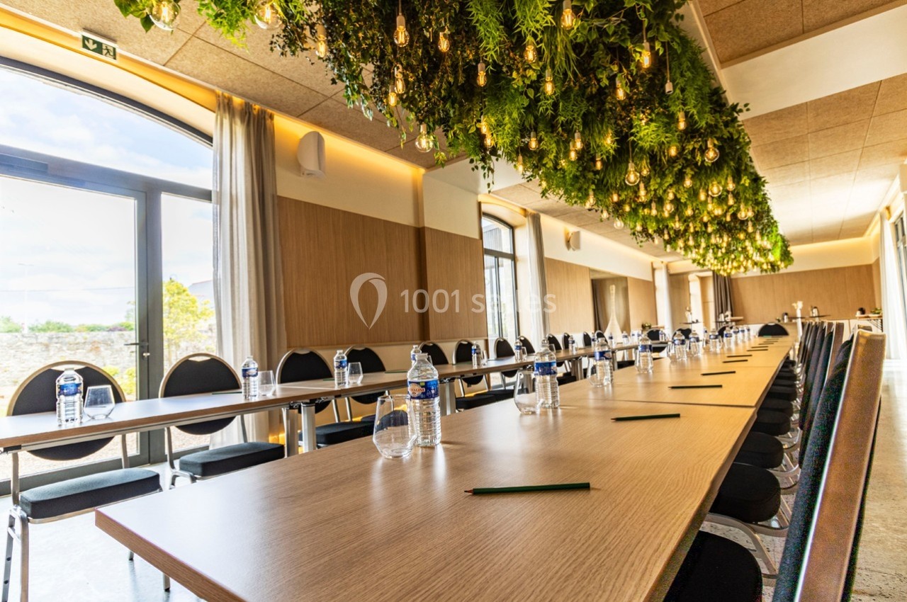 Salle de réunion lumineuse avec longues tables, chaises alignées, bouteilles d'eau et plafond décoré de plantes suspendues.