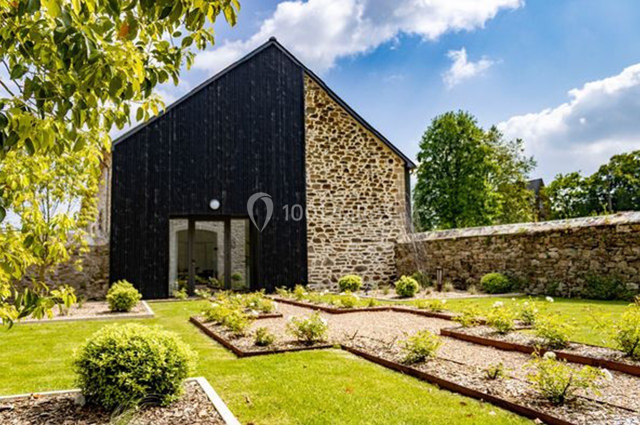 Façade d'une maison en pierre et bois noir, entourée d'un jardin aménagé avec des parterres et des allées gravillonnées.