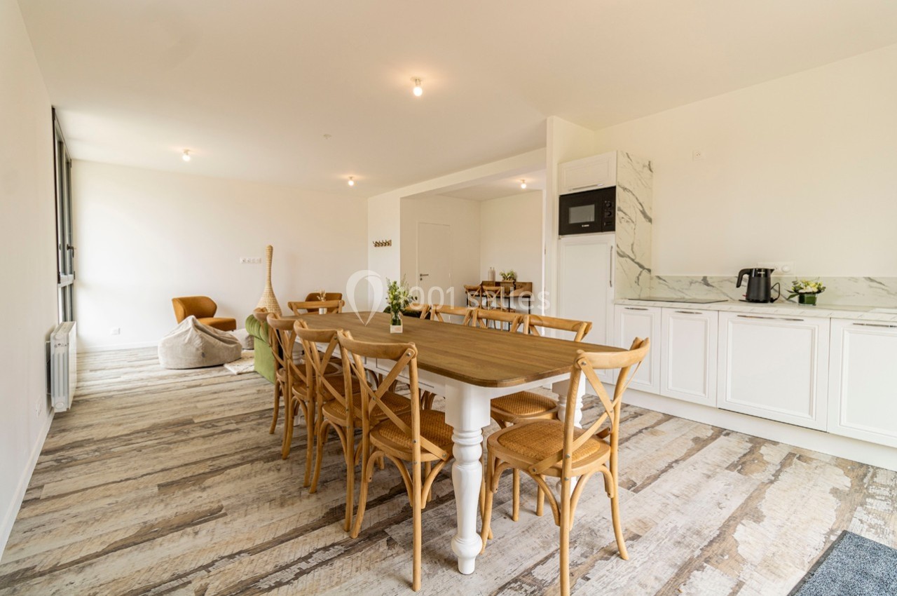 Salle à manger lumineuse avec une grande table en bois, chaises en bois clair et cuisine ouverte équipée en arrière-plan.
