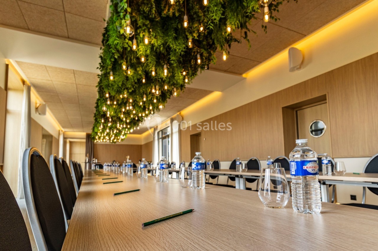 Salle de réunion avec une longue table en bois, des bouteilles d'eau, des verres, des stylos et un plafond décoré de plantes…