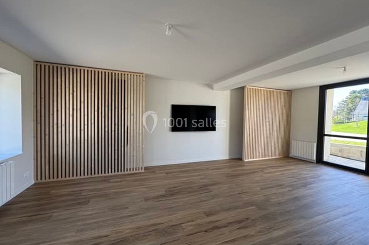 Salon vide avec parquet en bois, murs blancs, panneaux en bois clair, télévision murale et grande baie vitrée.