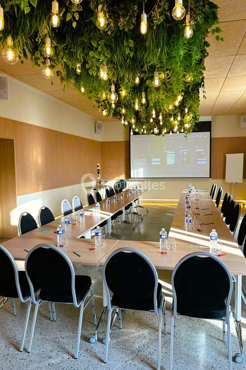 Salle de réunion lumineuse avec une grande table en U, chaises noires, écran de projection et plafond décoré de plantes…