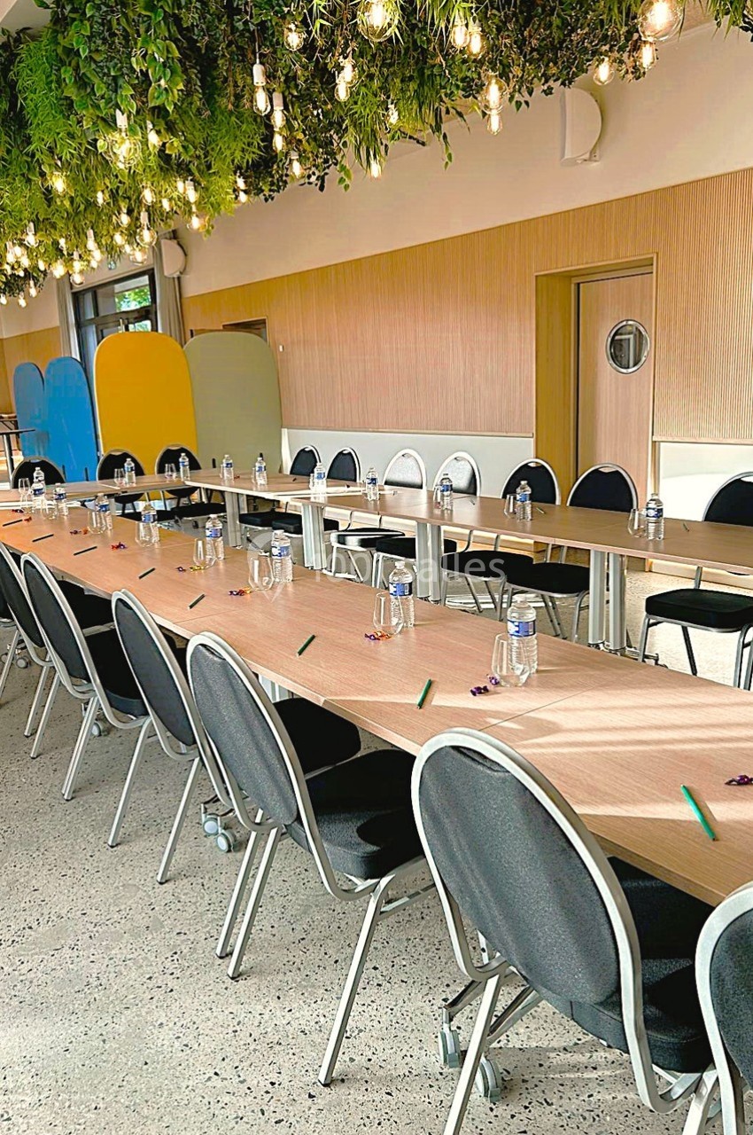 Salle de réunion avec longues tables alignées, chaises noires, bouteilles d'eau et crayons, sous un plafond végétalisé.