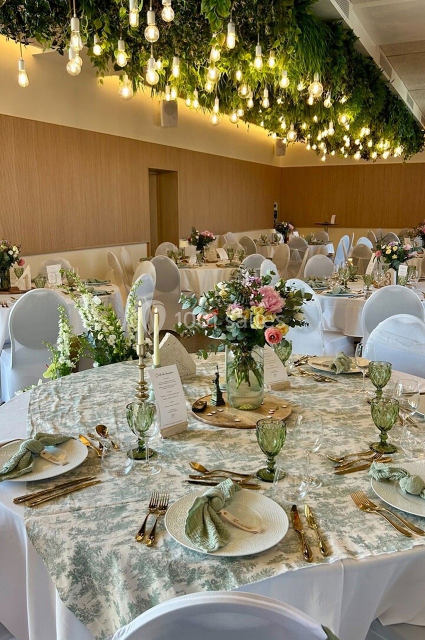 Salle de réception décorée avec des tables élégantes, nappes fleuries, vaisselle raffinée et plafond végétalisé éclairé.