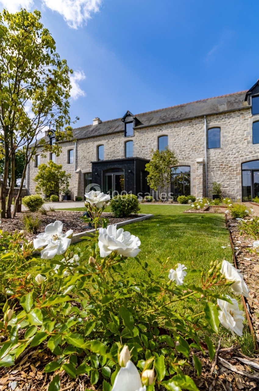 Façade d'une maison en pierre entourée de verdure, avec des roses blanches au premier plan et un ciel bleu dégagé.