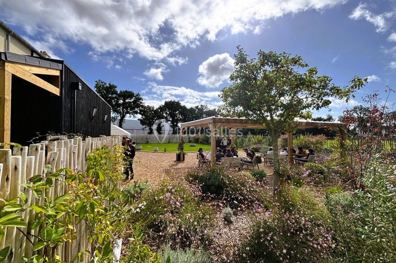 Jardin aménagé avec des plantes, une pergola en bois et des personnes assises sous un ciel partiellement nuageux.