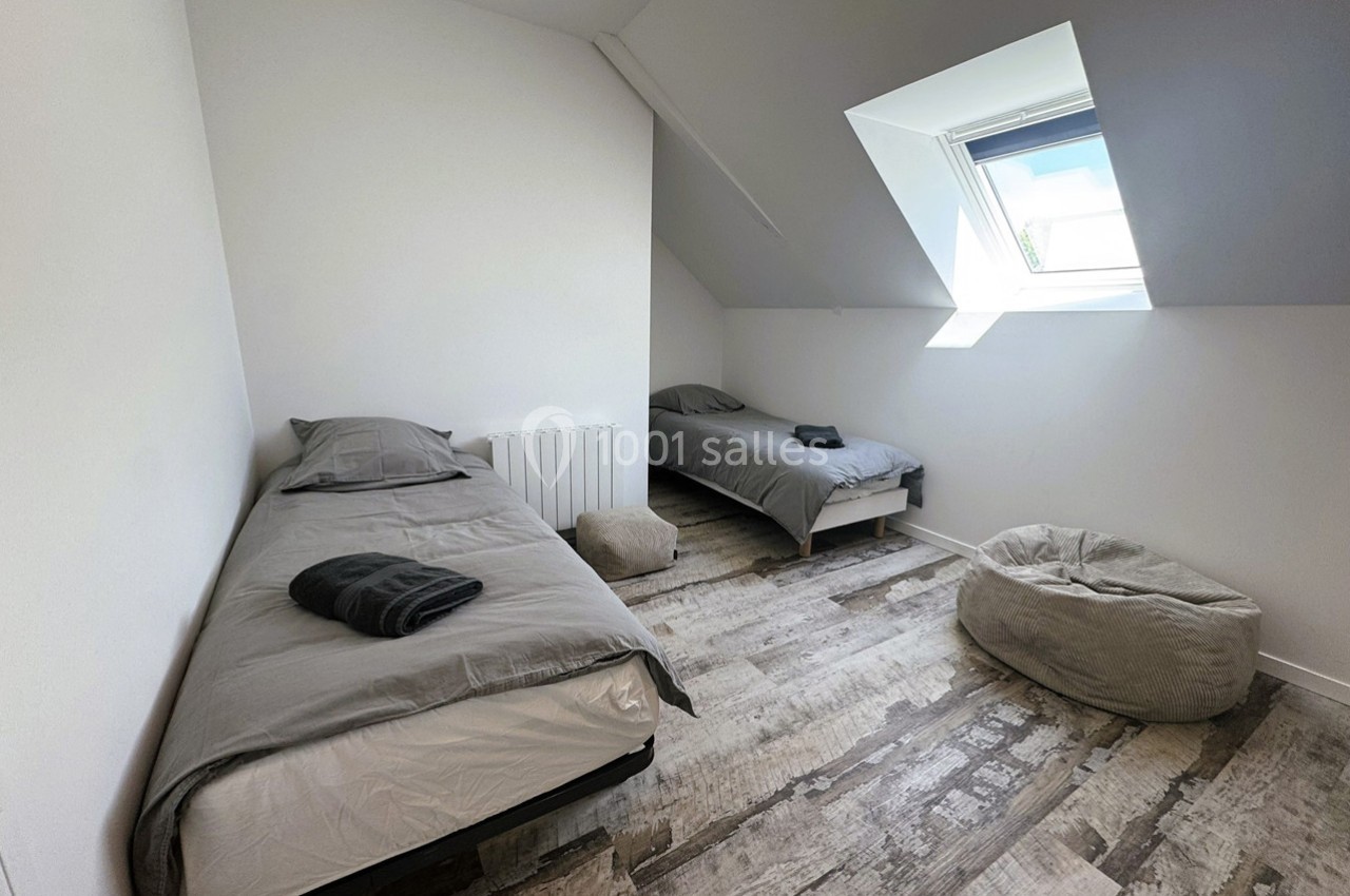 Deux lits simples avec draps gris dans une chambre lumineuse sous les combles, avec un pouf et un sol en bois clair.