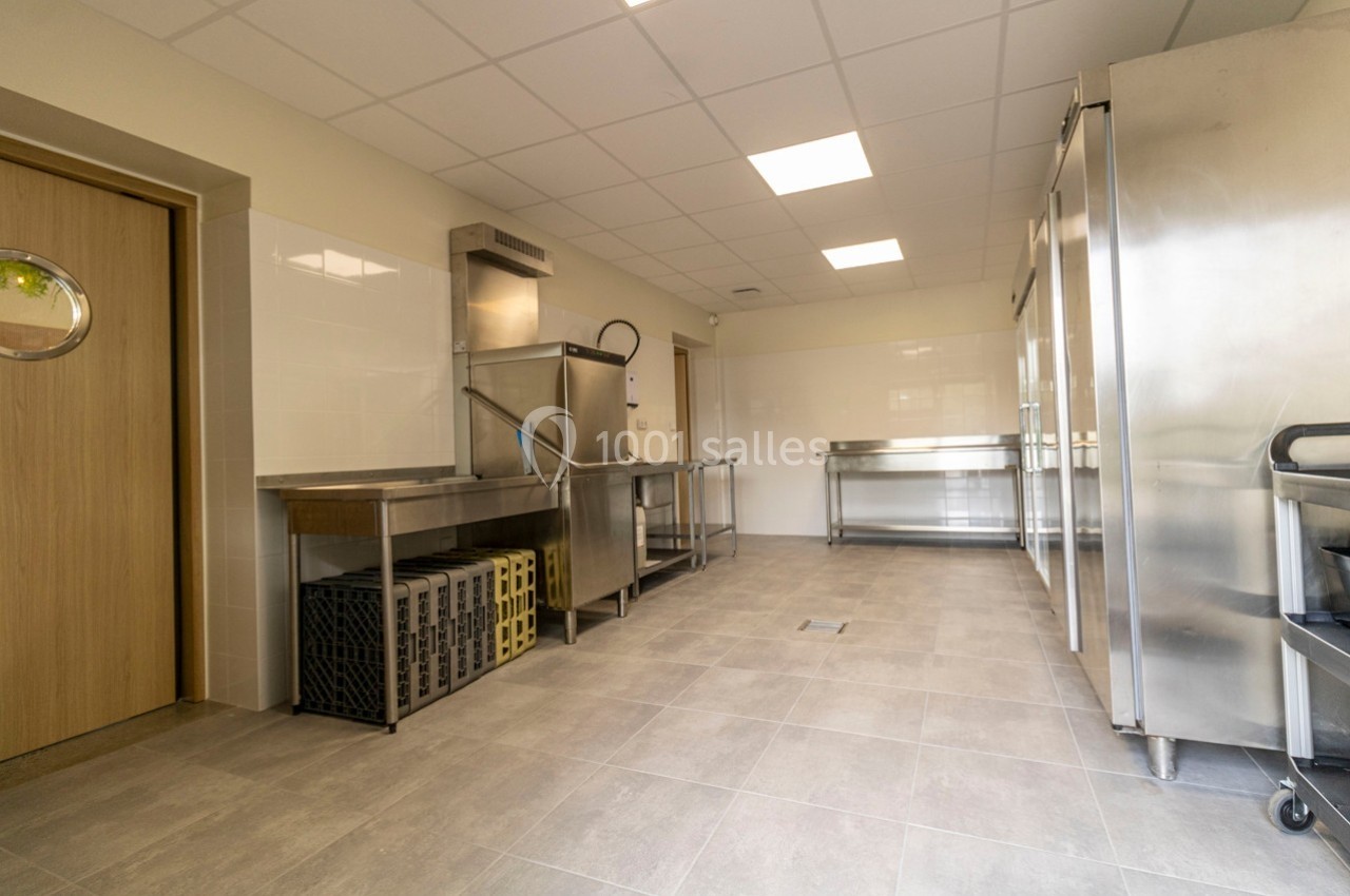 Cuisine professionnelle avec équipements en inox, lave-vaisselle industriel, étagères et sol en carrelage gris.