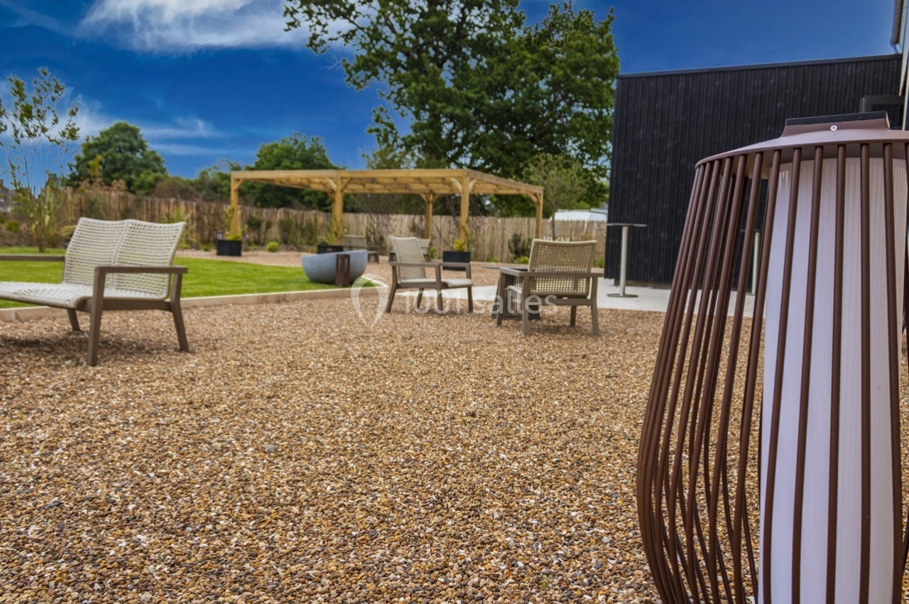 Cour extérieure avec gravier, mobilier de jardin, pergola en bois et végétation en arrière-plan sous un ciel bleu.