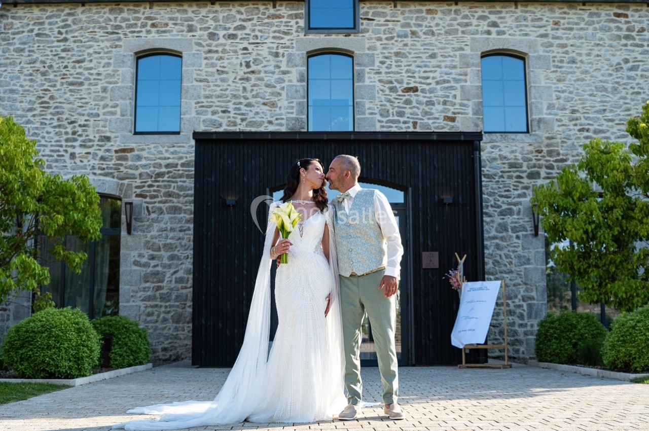 Un couple en tenue de mariage pose devant un bâtiment en pierre avec des arbres et une pancarte en arrière-plan.