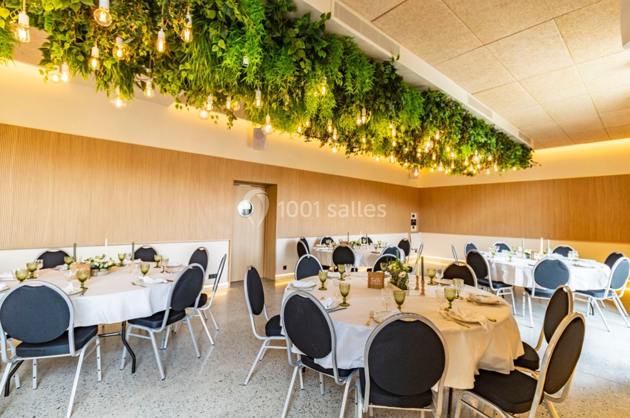 Salle de réception décorée avec des tables rondes dressées et un plafond orné de plantes suspendues et de lumières.