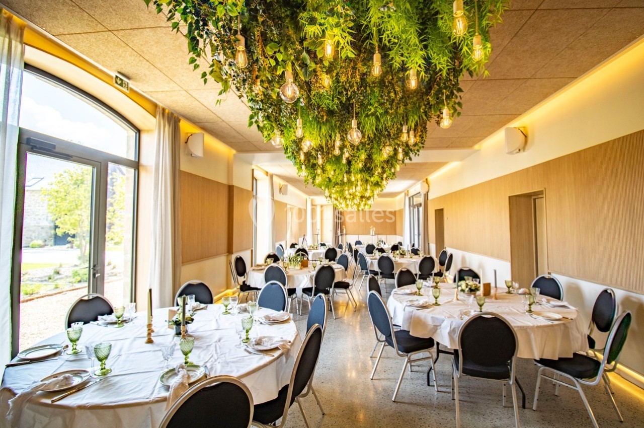 Salle de réception lumineuse avec tables rondes dressées et un plafond décoré de végétation et d'ampoules suspendues.