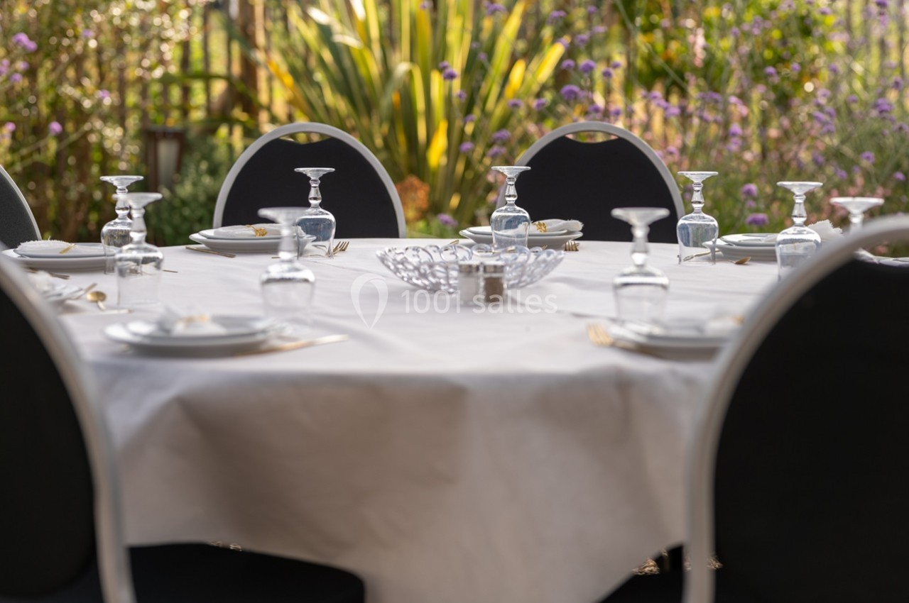 Table ronde dressée avec nappe blanche, assiettes, verres et couverts, entourée de chaises noires dans un jardin fleuri.