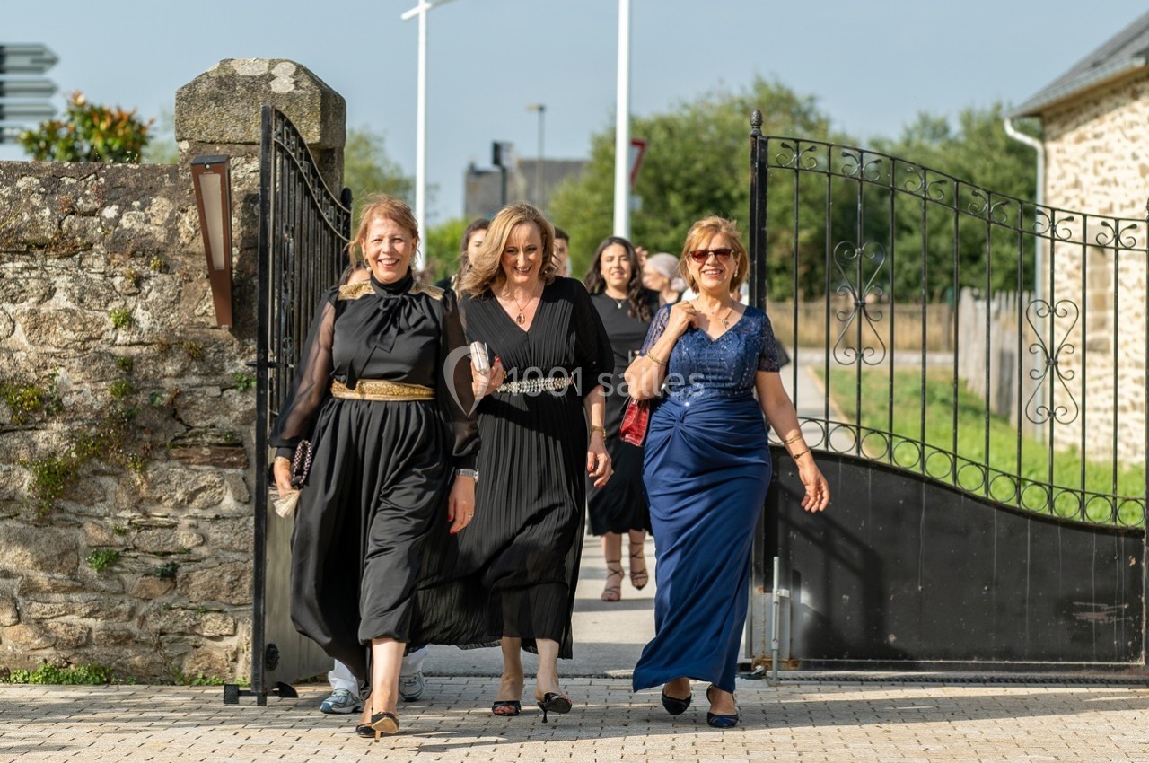 Des femmes élégamment vêtues marchent en groupe à l'extérieur, passant par un portail ouvert dans un cadre rural.