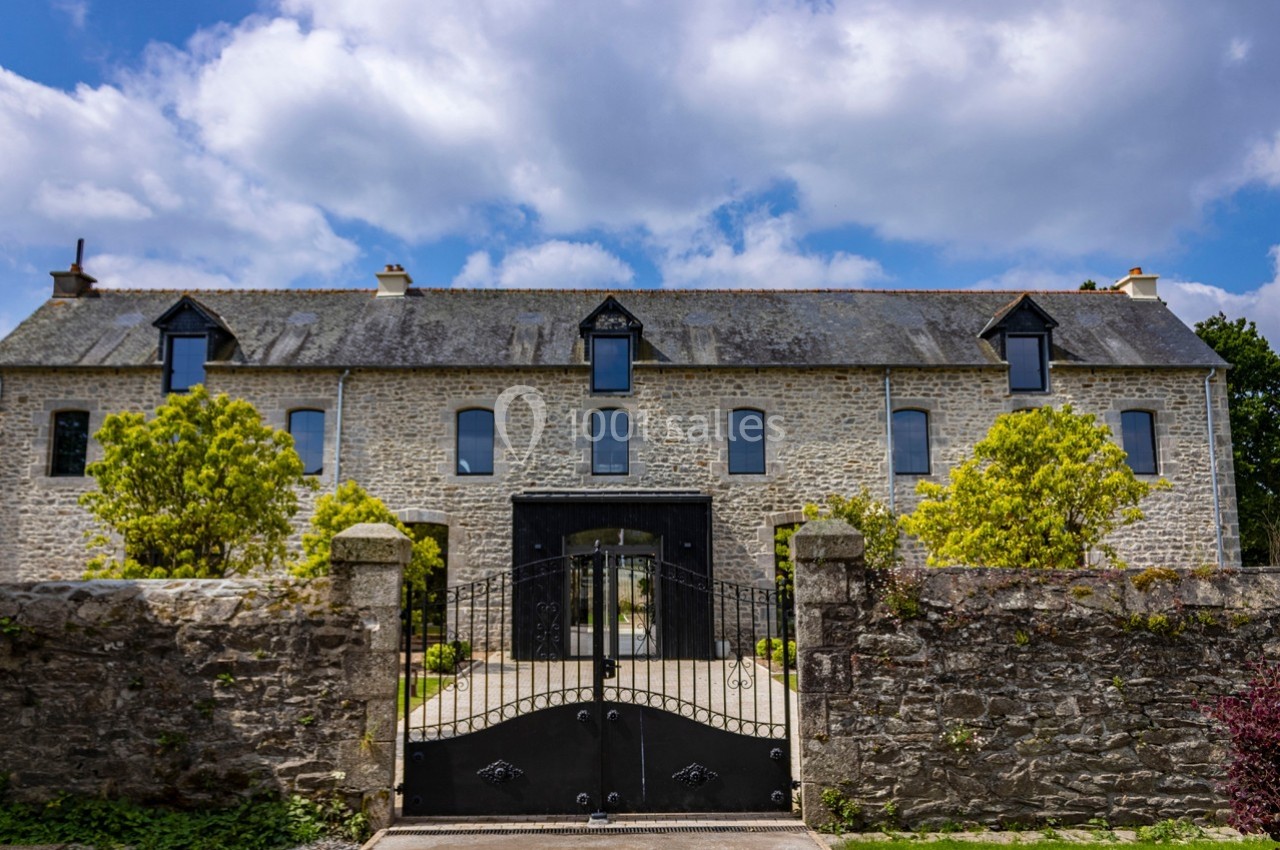 Façade d'une maison en pierre avec toit en ardoise, portail en fer forgé et arbres dans une cour clôturée.