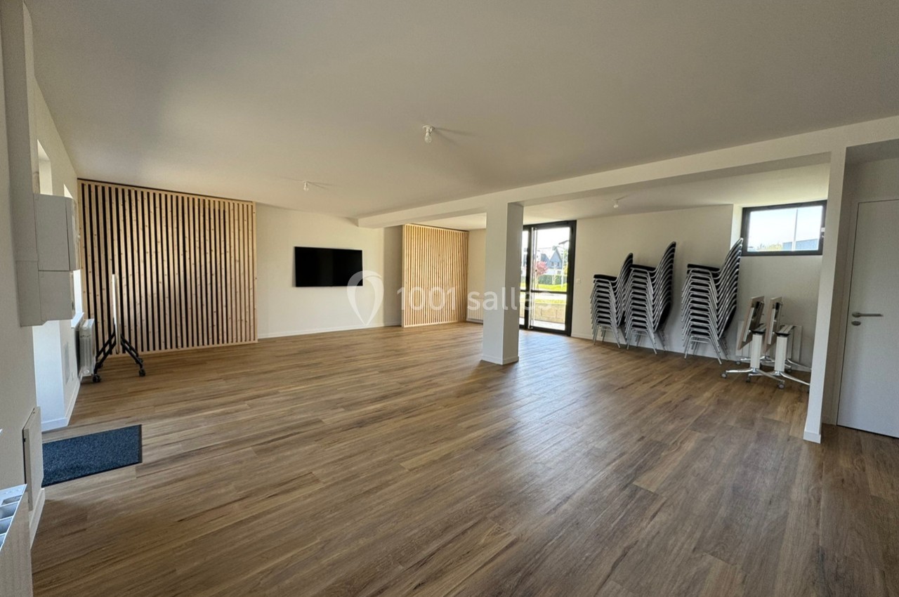 Salle lumineuse avec parquet, murs blancs, écran mural, chaises empilées et grandes fenêtres laissant entrer la lumière…