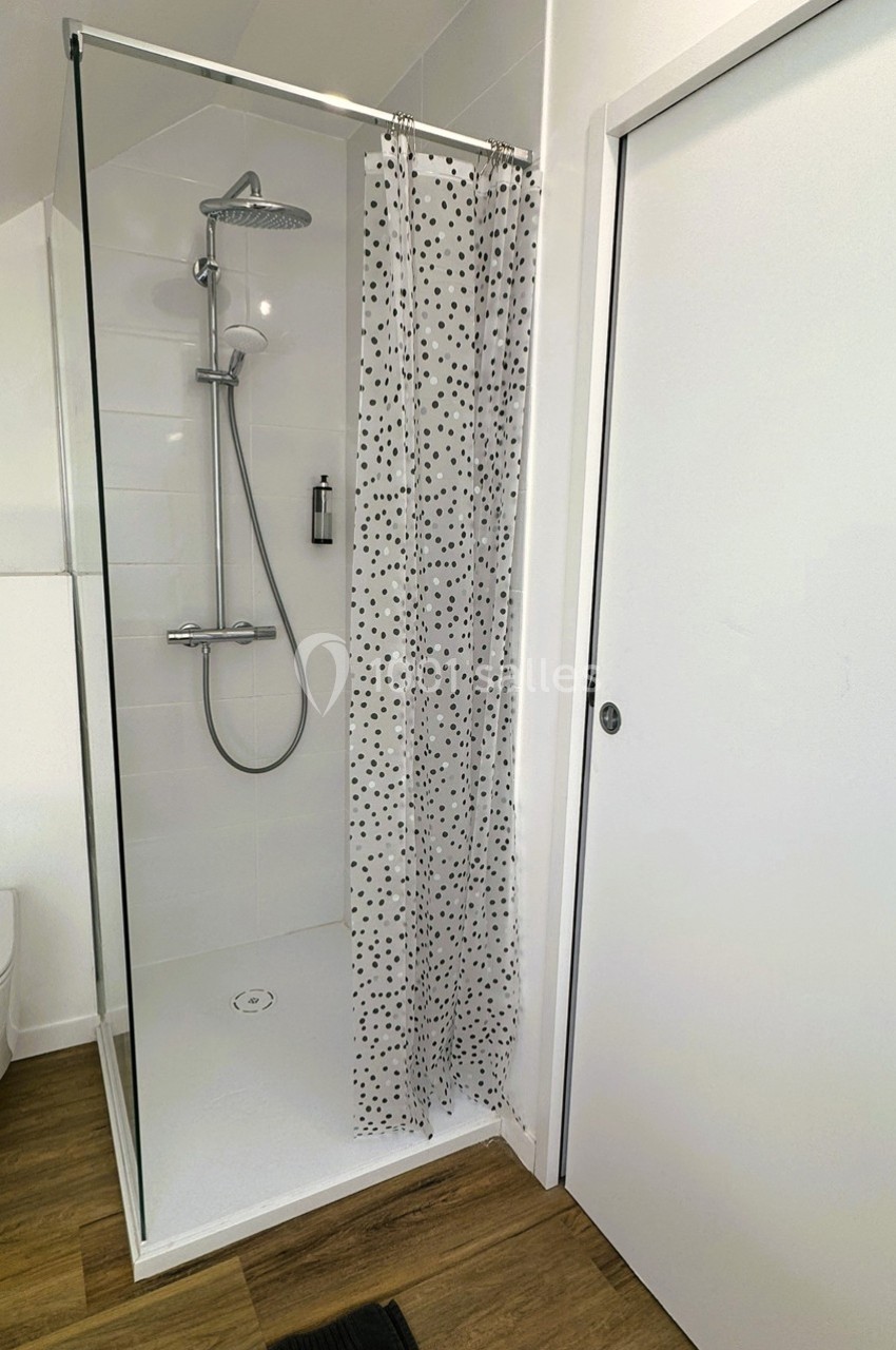 Douche avec paroi vitrée, rideau à motifs noirs, pommeau de douche mural et sol en bois clair.