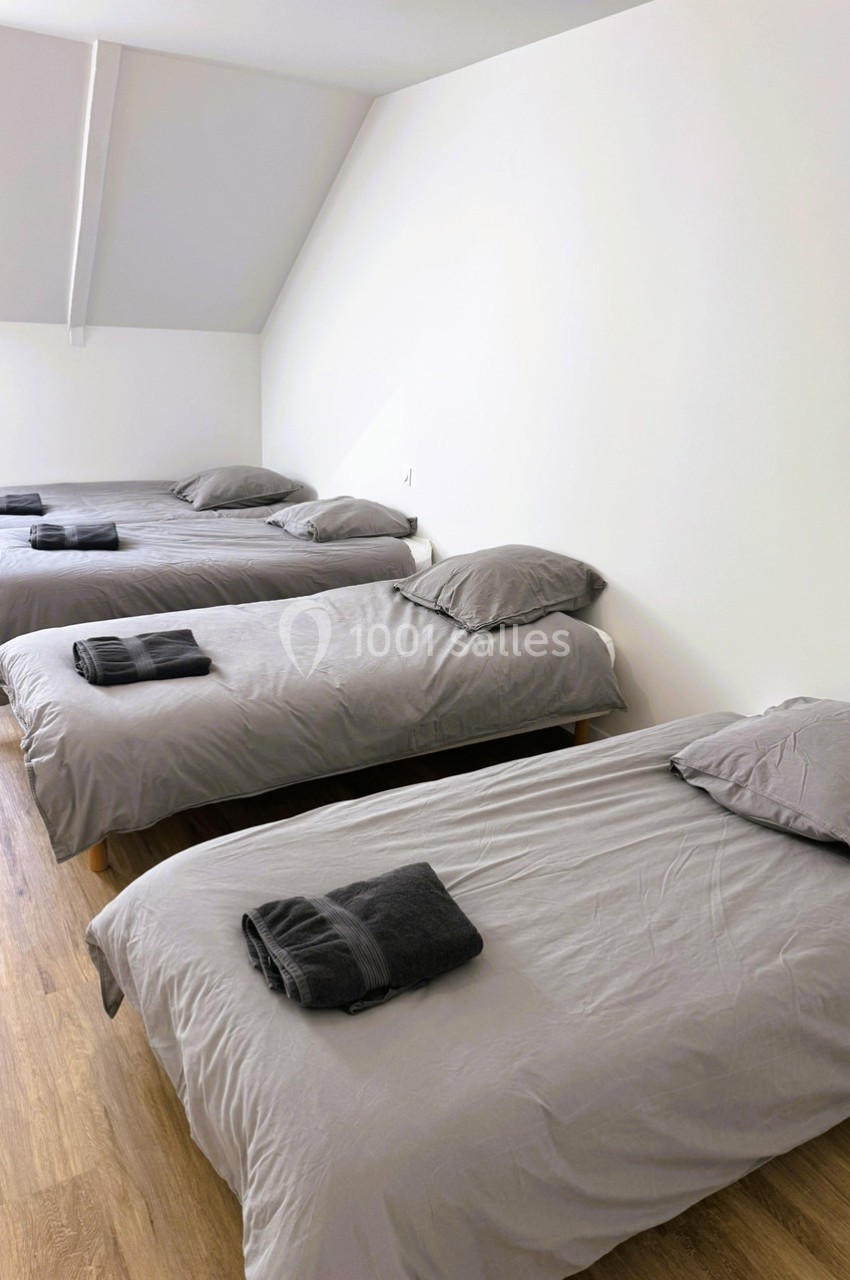 Trois lits simples avec draps gris et serviettes pliées sur un sol en parquet dans une pièce lumineuse.
