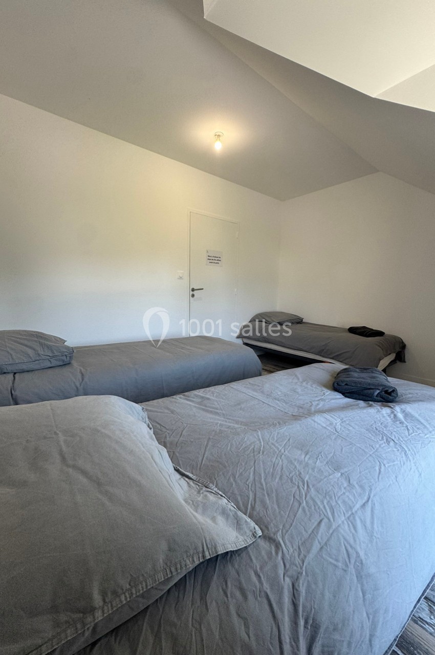Chambre avec trois lits simples, draps gris et lumière naturelle entrant par une fenêtre hors champ.