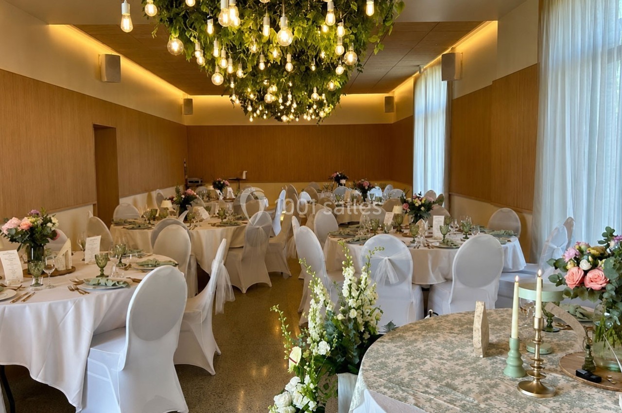 Salle de réception décorée pour un événement, avec tables rondes dressées, nappes blanches et lustre végétal suspendu.