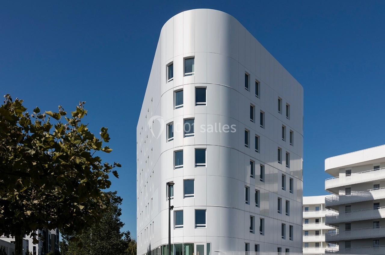 Bâtiment moderne blanc à plusieurs étages avec des fenêtres rectangulaires, entouré d'arbres et d'autres structures.