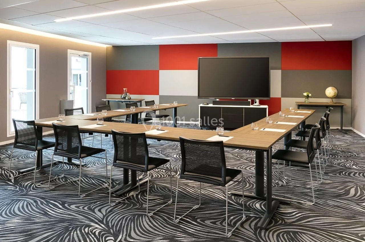 Salle de réunion moderne avec tables en U, chaises noires, écran large et décoration murale colorée.